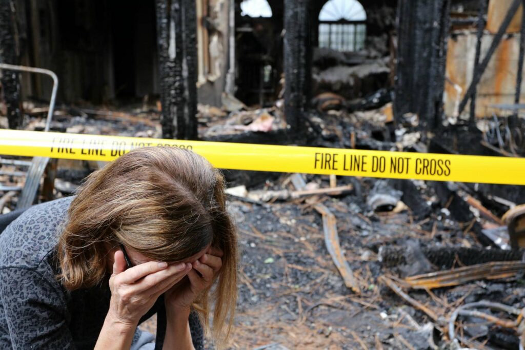 A woman covers her face in distress near a fire-damaged building behind yellow "Fire Line Do Not Cross" tape.