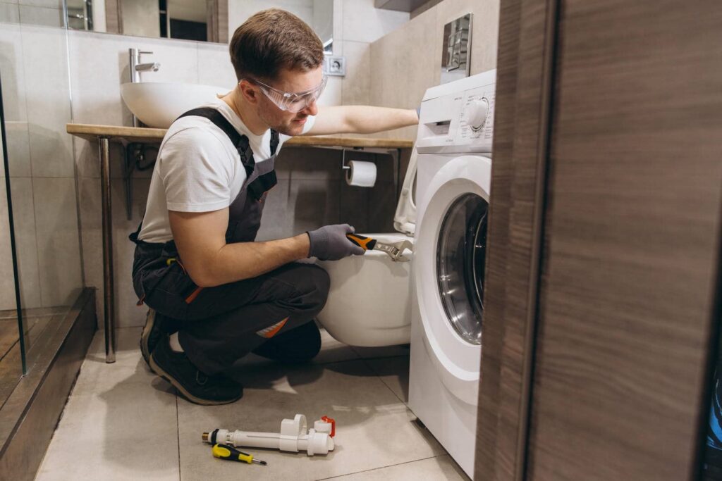 A man wearing safety glasses and gloves repairs a washing machine in a bathroom.