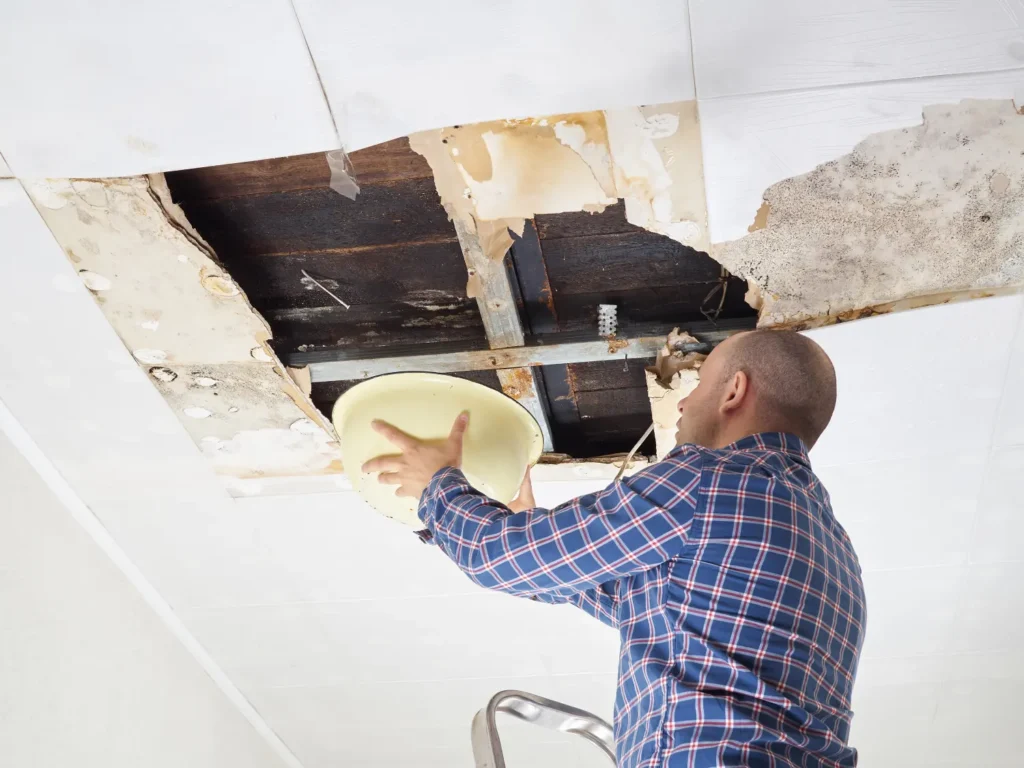 Man on ladder inspecting water-damaged ceiling with large hole and mold.