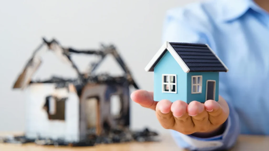 A hand holding a small intact blue house model with a burned, destroyed house in the background.