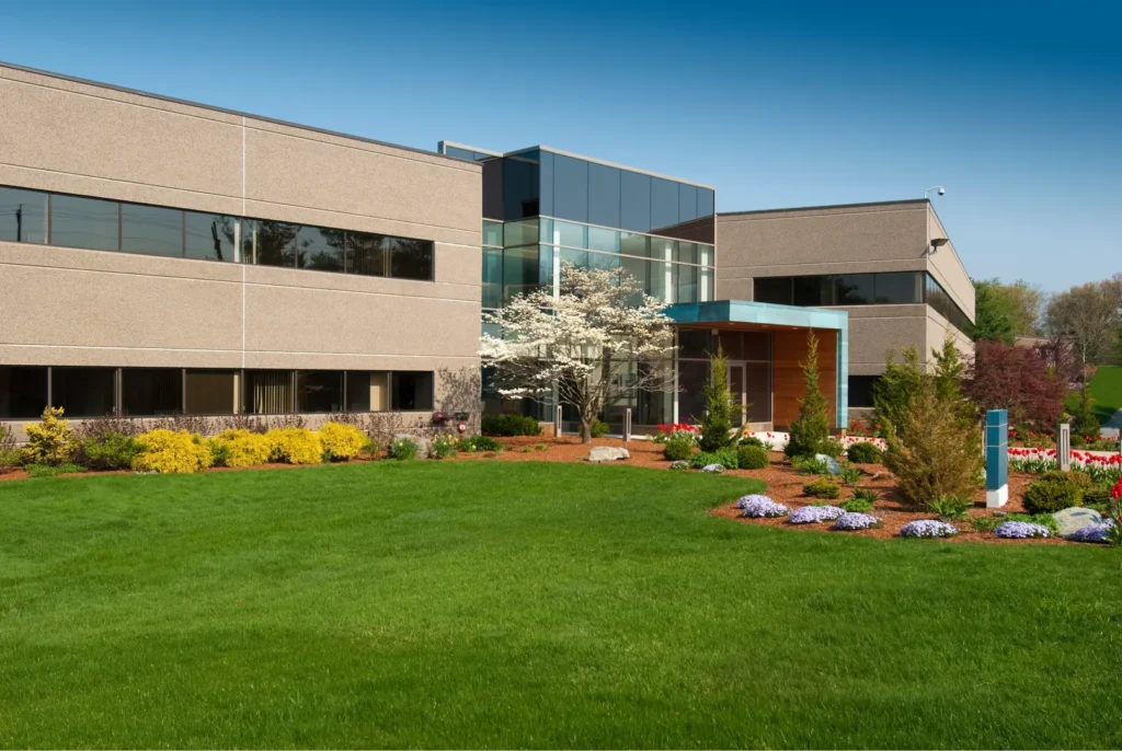 Modern office building with large windows surrounded by green lawn and landscaped garden beds.