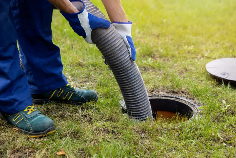 Worker wearing gloves inserts a flexible hose into an open septic tank on grass.