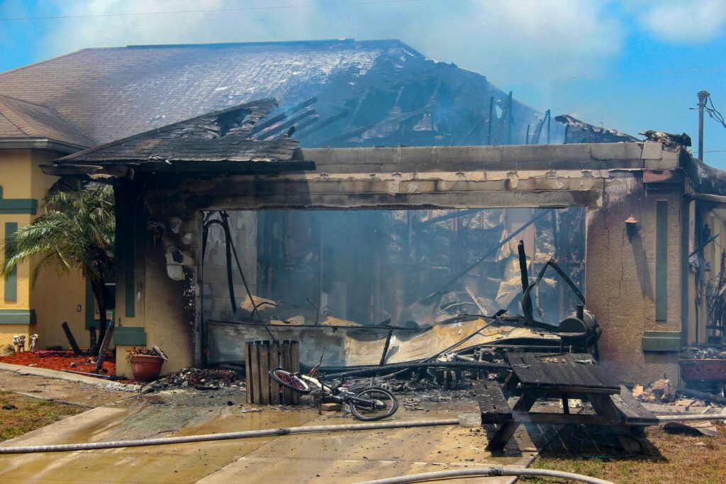 Burned house with collapsed garage and charred debris including a fallen bicycle and picnic table.