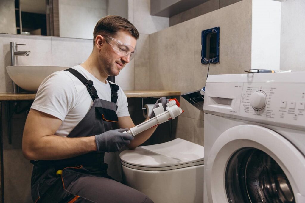 Man wearing safety glasses and gloves holding a plumbing part while kneeling next to a toilet and washing machine.