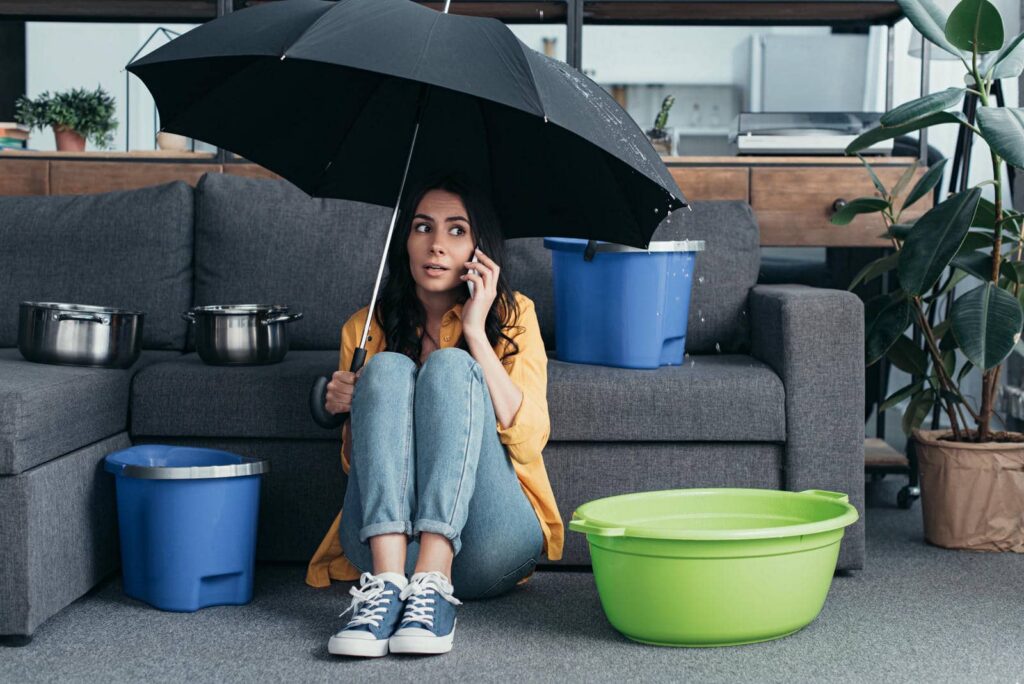Woman sitting on floor under umbrella indoors with buckets catching water from a leak.