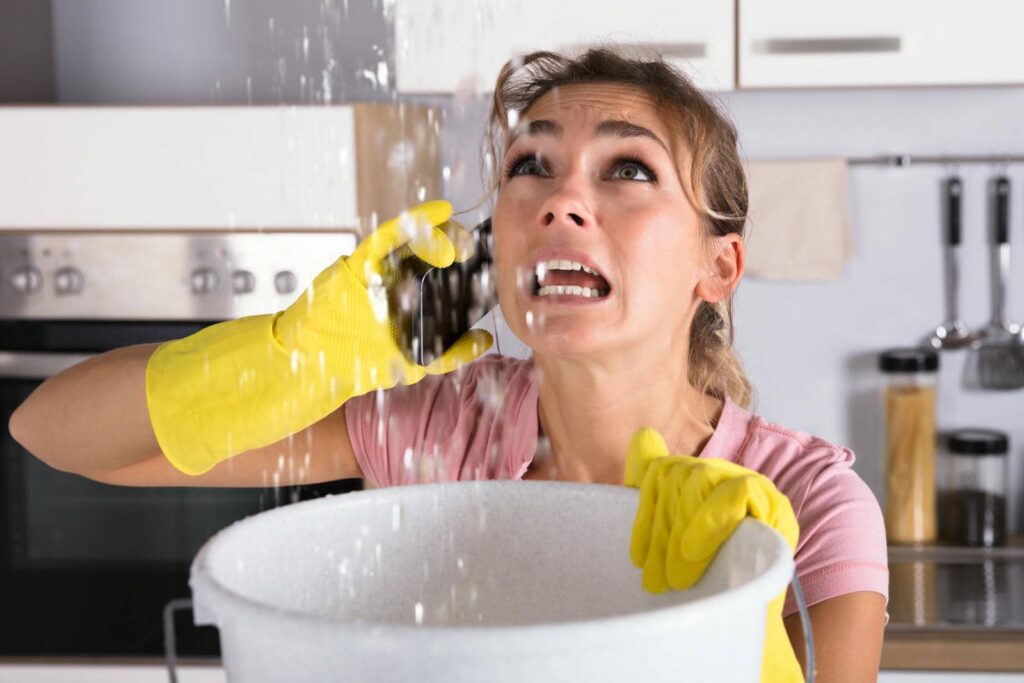Woman in yellow gloves holding a bucket and looking distressed as water leaks above her in a kitchen.